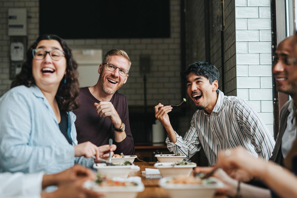 En grupp multietniska kollegor sitter vid ett bord på en restaurang i Göteborg, Sverige, och delar en gemensam lunchrast fylld av skratt och glädje